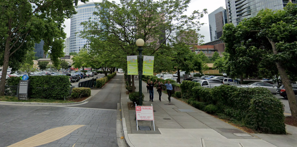 A photo of a sidewalk and narrow street running between two surface level parking lots. 