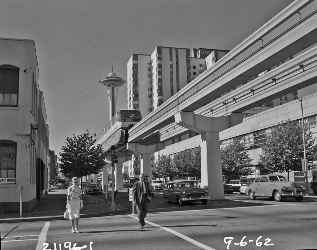 A black and white photograph of a monorail train running above a street in Downtown Seattle with a man and woman crossing beneath on foot at a crosswalk. 