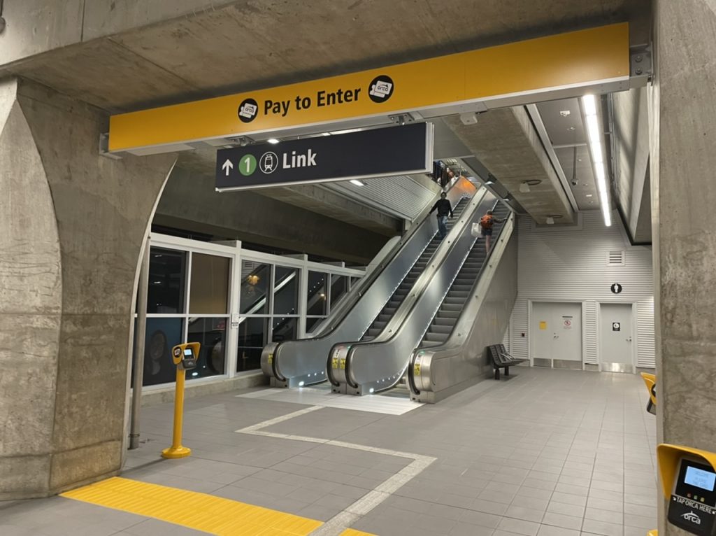 A photo showing the entrance to a light rail station with a yellow sign stating pay to enter and escalators appearing behind it. 
