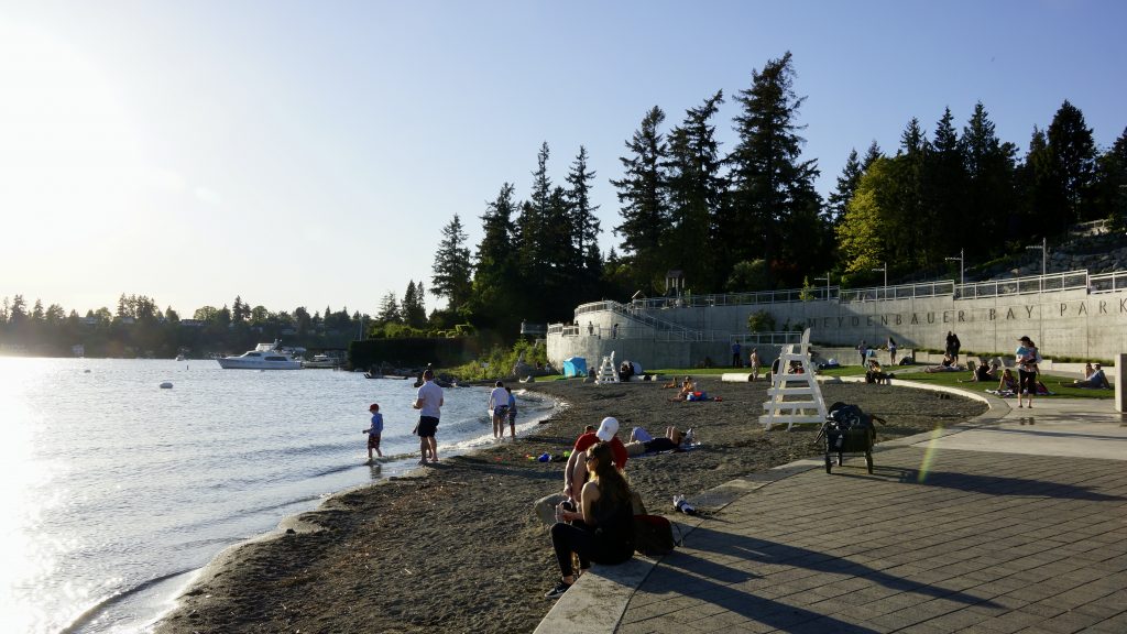 A photo of people sitting a small beach on a sunny day with evergreen trees in the background.