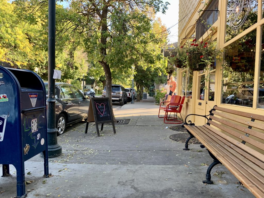 A photo of a sidewalk on a small commercial street with a post office box and bench.