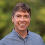 A photo shows a White man with short brown hair wearing a button down blue shirt.