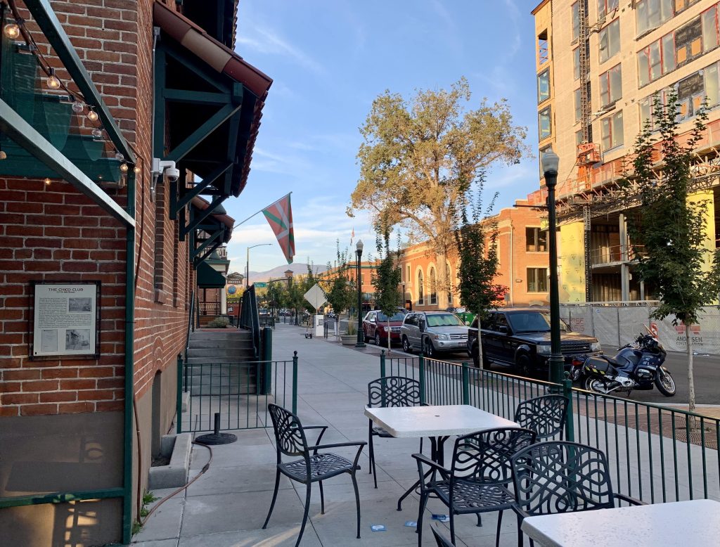 A photo shows an outdoor cafe with tables and chairs. A Basque flag is hung on the building. Across the street a new taller building is under construction. 