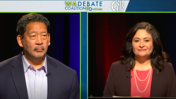 A photo shows a man with greying black hair and mustache on the left side of the screen and a woman with shoulder length black hair and a red shirt beneath jacket on the right side of the stage.