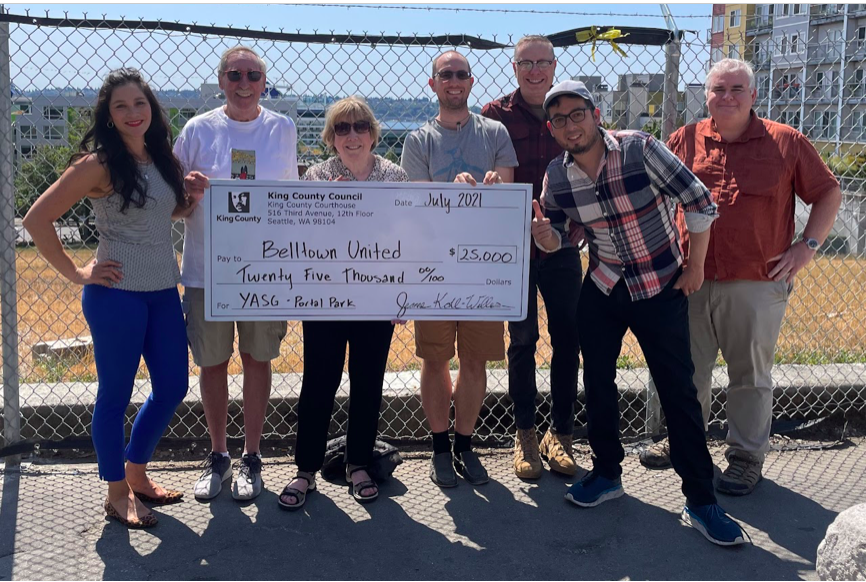 A photo show seven adults standing in front of a chainlink fence holding a huge check for $25,000 awarded to Belltown United by the King County Council. 