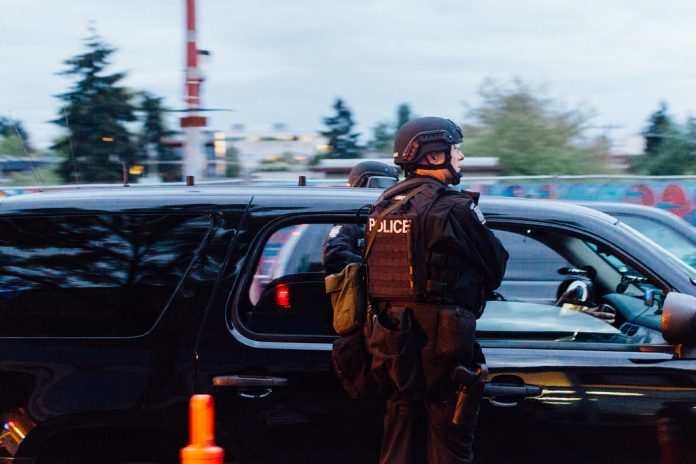 A photo shows a police officer in riot gear standing next to a patrol car.