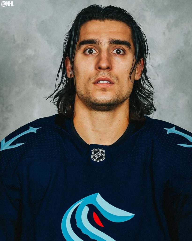 A man with long dark hair wearing a Seattle Kraken hockey jersey stares dazed at the camera. 