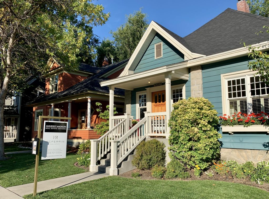 A photo showing a blue bungalow style house with a for sale sign in the front yard.