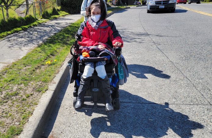 wheelchair user in road A woman wearing a red jacket and face mask rides her power wheelchair in the road with a person walking behind her and a car approaching.