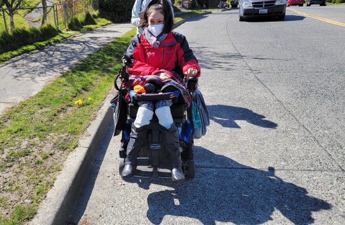 A woman wearing a red jacket and face mask rides her power wheelchair in the road with a person walking behind her and a car approaching.