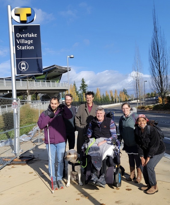 A photo shows a man standing in a purple coat with a white cane, near to two men standing in grey coats behind a man seated in a wheelchair with a seeing eye dog. Two women also in coats are crouching next to him.