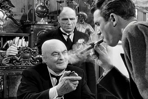 A black and white photo of a man puffing on a cigar with two older men in suits standing before him. 