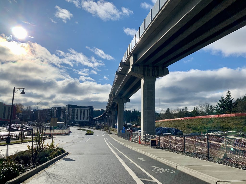 A photo of elevated light rail tracks and a road with a bike lane leading to mixed use apartment buildings. 