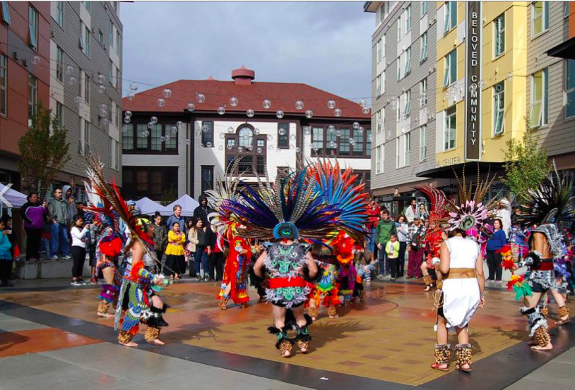 A photo of dancers in brightly colored costumes with featured headdresses dancing in a plaza surrounded by buildings. 
