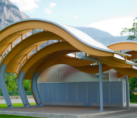 Lecture Series Spotlights Indigenous Architecture A photo of a wave shaped outdoor pavilion made from wood in front of mountains.