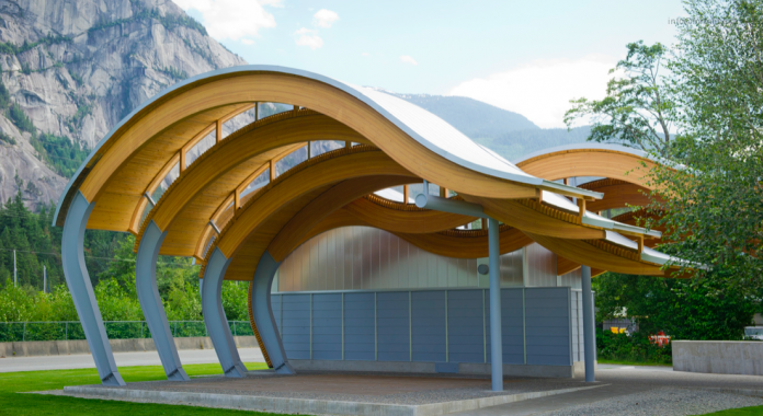 A photo of a wave shaped outdoor pavilion made from wood in front of mountains.
