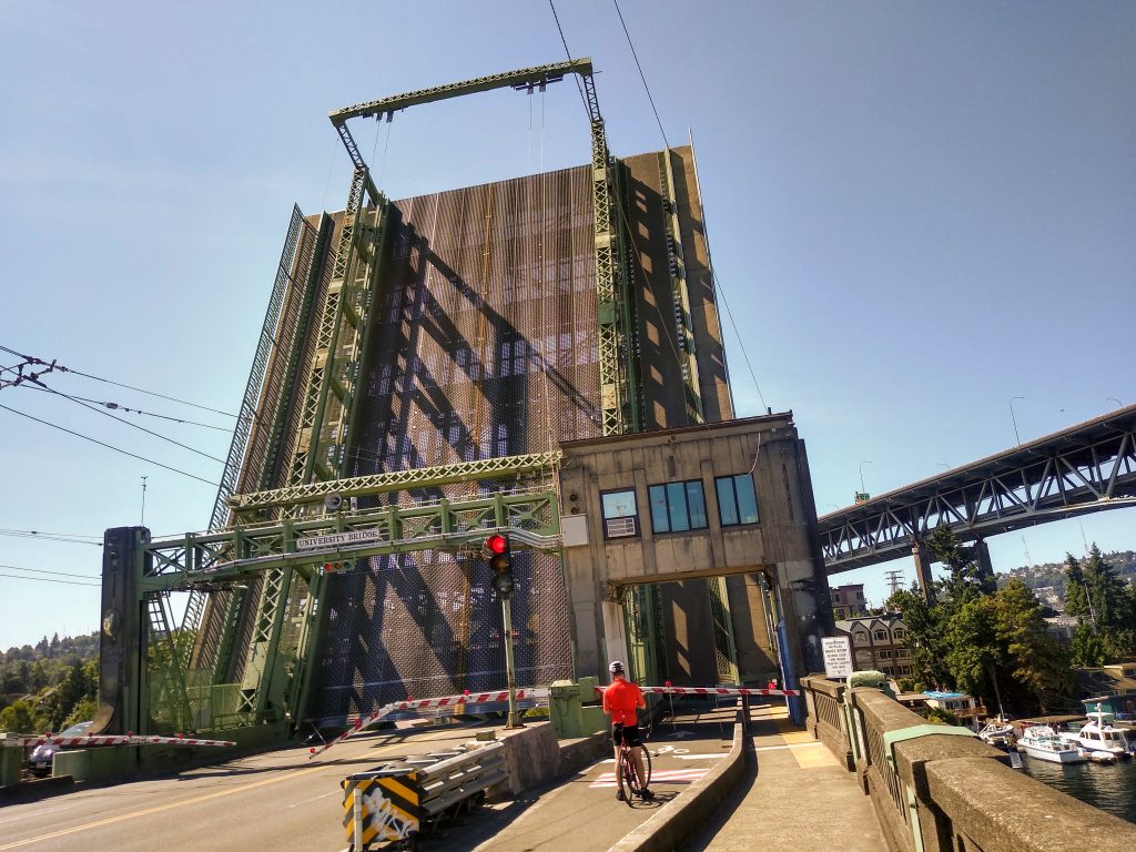 A photo of a raised drawbridge with a cyclists waiting in front of it.