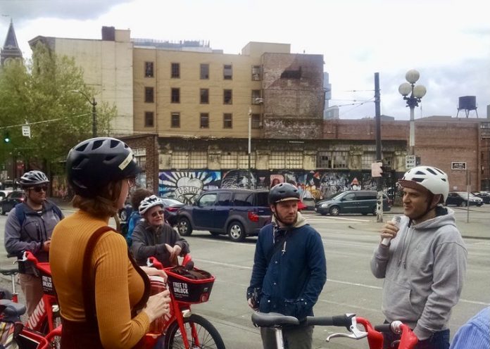 A group of people wearing bike helmets and standing near bikes listen to a man speaking.