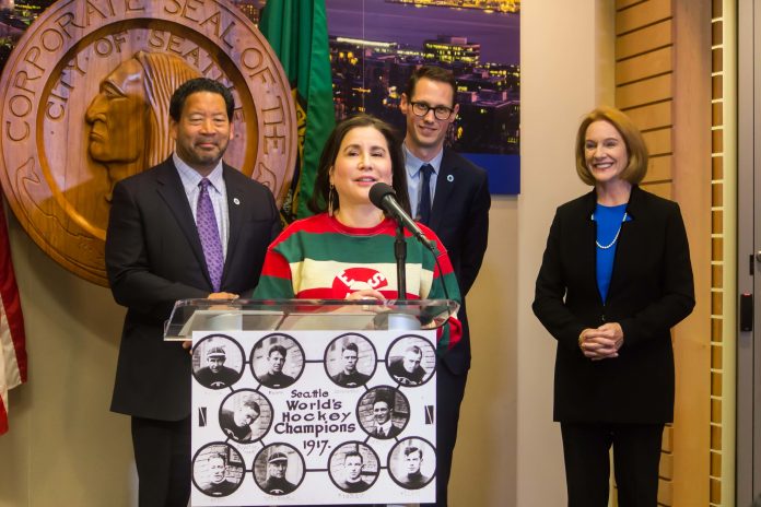 Debora Juarez at NHL announcement Juarez speaks at a podium with a hockey jersey with the City Seal in the background.
