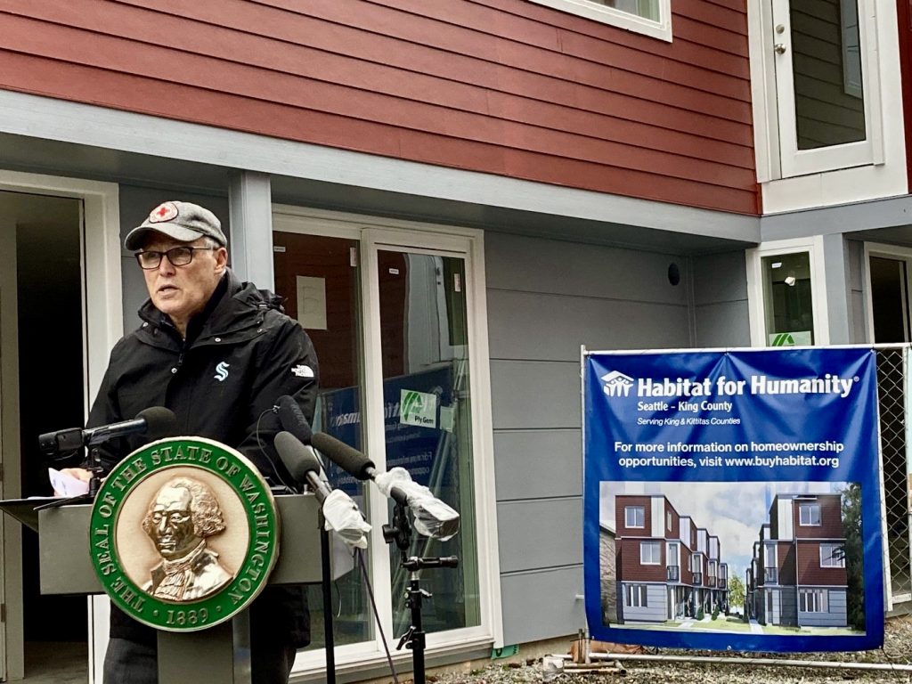 A photo of a man speaking at a podium decorated with the seal of the State of Washington in front of a townhouse and a sign for habitat for humanity.