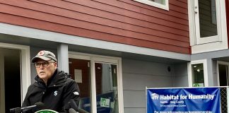 A photo of a man speaking at a podium decorated with the seal of the State of Washington in front of a townhouse and a sign for habitat for humanity.