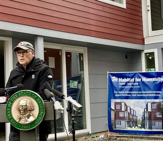 Governor Inslee Backs Fourplex Zoning and $815 Million Push to Reduce Homelessness A photo of a man speaking at a podium decorated with the seal of the State of Washington in front of a townhouse and a sign for habitat for humanity.