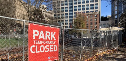 A photo of a chainlink fence and a sign reading "park closed" in front a open grassy area.