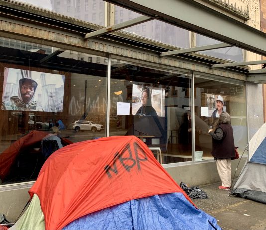 King County Estimates 40,800 Residents Experienced Homelessness in 2020 A photo of a tent in front of a glass window with exhibiting large portrait style photos of people.