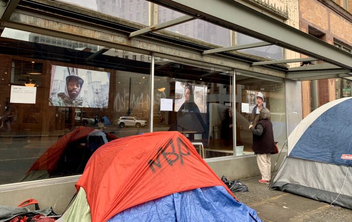 Homeless Photograph Exhibit in Pioneer Square A photo of a tent in front of a glass window with exhibiting large portrait style photos of people.