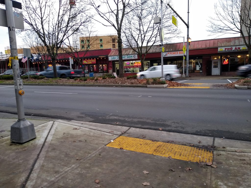 A wide street with a median, with a tactile strip on either side and in the median. The near side of the street has no markings. The far side has two faint lines.