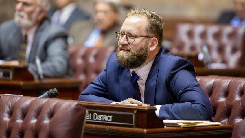 Image description: Liias: a light-haired man in glasses and a beard in a blue suit with his hands clasped sitting in a leather chair in senate chambers.