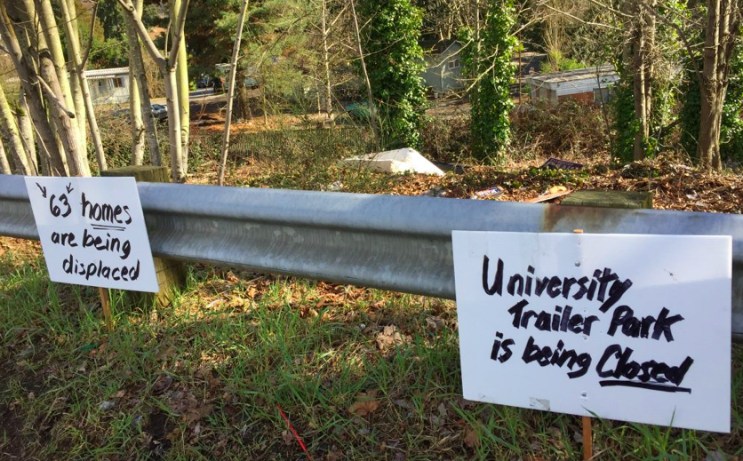A photo of a guard rail with signs reading "63 homes are being displaced" and University Trailer Park is being closed."
