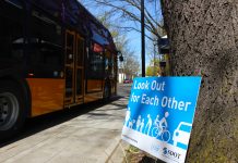 As Statewide Traffic Safety Emergency Escalates, Seattle’s Vision Zero Department Charts New Path A blue sign leans against and tree and it reads "Look out for reach other" with an image of people walking, biking, and rolling next to a car. A Metro bus is in the background.