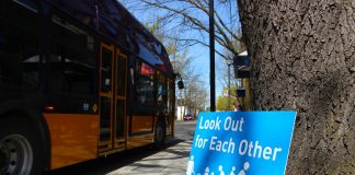 A blue sign leans against and tree and it reads "Look out for reach other" with an image of people walking, biking, and rolling next to a car. A Metro bus is in the background.