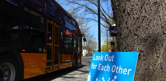 A blue sign leans against and tree and it reads "Look out for reach other" with an image of people walking, biking, and rolling next to a car. A Metro bus is in the background.