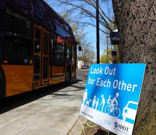 As Statewide Traffic Safety Emergency Escalates, Seattle’s Vision Zero Department Charts New Path A blue sign leans against and tree and it reads "Look out for reach other" with an image of people walking, biking, and rolling next to a car. A Metro bus is in the background.