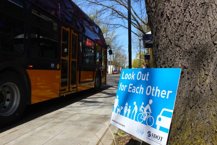 Look out for each other A blue sign leans against and tree and it reads "Look out for reach other" with an image of people walking, biking, and rolling next to a car. A Metro bus is in the background.