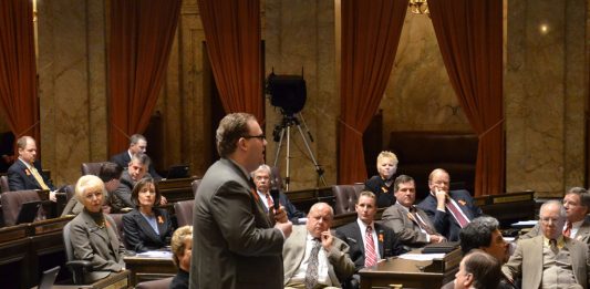 A photo of a man wearing a suit in a senate chamber standing and speaking while other people in suits sit and listen.