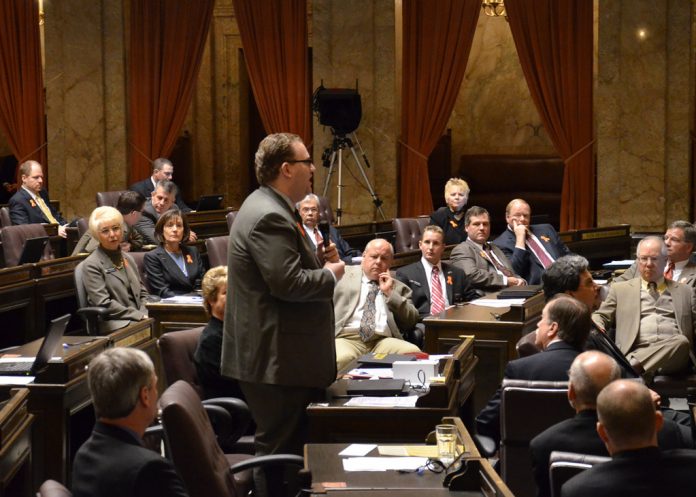 A photo of a man wearing a suit in a senate chamber standing and speaking while other people in suits sit and listen.