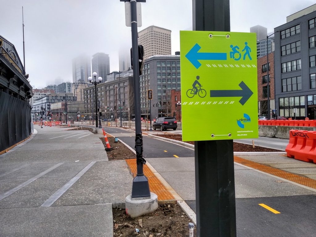 Bike trail and wide sidewalk with green sign, one arrow with wheelchair user and sidewalk user and another with bike user.