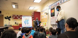 Children sit in a classroom and listen to a story being read aloud by a man in a suit wearing a mask.