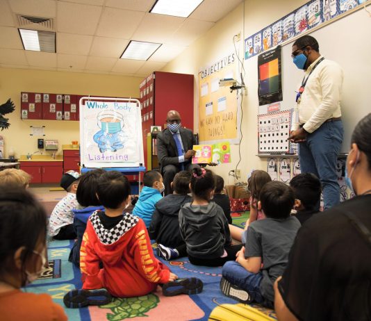 What Seattle Public Schools Needs to Say Right Now Children sit in a classroom and listen to a story being read aloud by a man in a suit wearing a mask.
