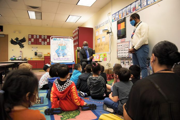 Children sit in a classroom and listen to a story being read aloud by a man in a suit wearing a mask.