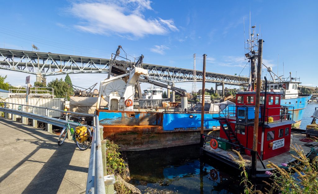 A photo of boats in the water next to a bike path with a bike parked on it. 