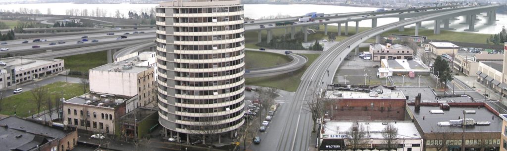 A looming elevated freeway structure with multiple multi-lane off ramps looms over city streets.