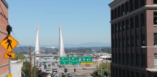 A photo of an crosswalk on a wide street with street signs and a bridge in the distance.