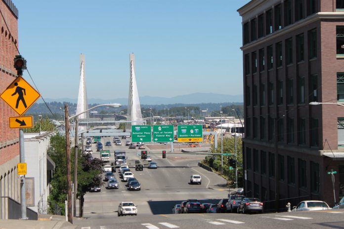 A photo of an crosswalk on a wide street with street signs and a bridge in the distance.