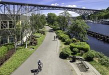 Seattle Street Ends: Fremont to the University Along Lake Union and Portage Bay A photo of a trail with a cyclist riding on it next to a canal with a bench overlooking it and a bridge in the distance.