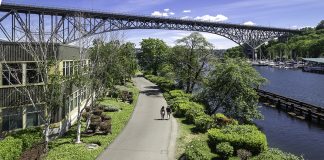 A photo of a trail with a cyclist riding on it next to a canal with a bench overlooking it and a bridge in the distance.