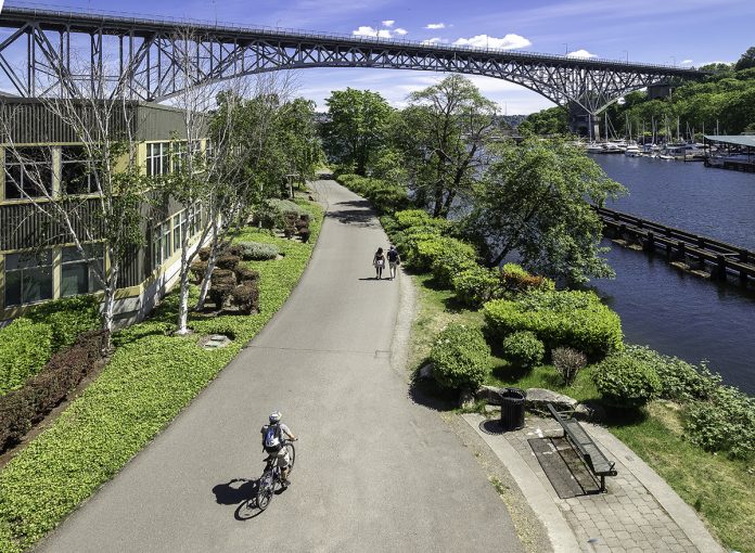 A photo of a trail with a cyclist riding on it next to a canal with a bench overlooking it and a bridge in the distance.